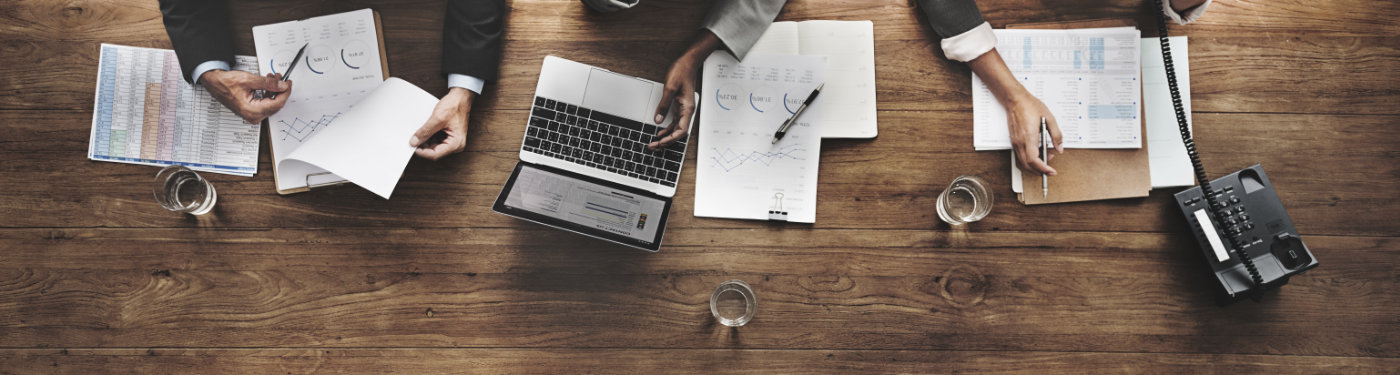 Rebecca Hughes | In-Company Training / Business English Banner Image | Large wooden table seen from above, 3 people working with pen and paper, one is on the phone and one is using a laptop